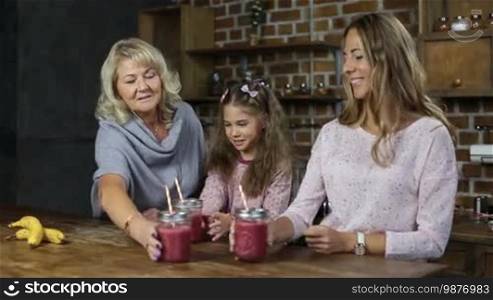 Portrait of three-generation family toasting red berry smoothies in mason jars and smiling over a modern kitchen background. Beautiful grandmother, daughter, and little granddaughter holding fresh berry smoothie and looking at the camera with toothy smiles.