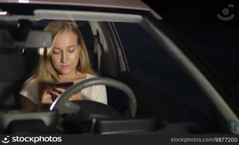 Positive beautiful long blonde hair female sitting in driver's seat and browsing the net with mobile phone on a summer night. Front view. Charming young woman in modern car using smart phone at night and smiling.