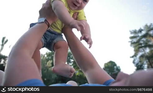 Positive father lying on his back, raising smiling infant son up in the air over beautiful landscape background. Closeup. Low angle view. Smiling toddler baby boy laughing while playing with father in park.