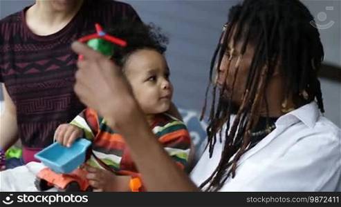Positive handsome African American dad with dreadlocks playing with his mixed race toddler child at home. Playful father having fun with his boy and playing with toys while little curly kid sitting on his lap and smiling closeup.