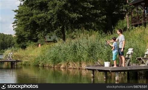 Positive hipster father teaching teenage boy how to fish with spinning rod on freshwater pond while enjoying weekend together on sunny summer day. Happy son failed to cast fishing rod from wooden pier on lake as he learns angling with dad's help.
