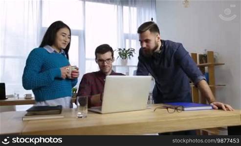 Positive hipster freelancers gathered around laptop PC in office to discuss new startup business. Multi-ethnic casual business team sharing ideas and brainstorming at workplace in modern office. Dolly