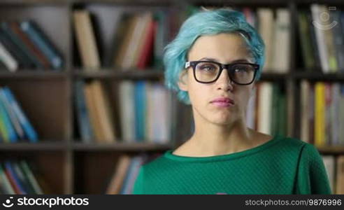 Positive human facial expressions and emotions. Portrait of an attractive hipster girl with blue hair wearing trendy eyewear looking and smiling broadly at the camera. Charming female college student in eyeglasses with a radiant smile posing at the library.