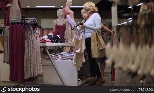 Positive senior female friends choosing new clothes in shop. Beautiful blonde women with apparels in their hands shopping trendy clothes in boutique in shopping mall. Cheerful ladies buying garments during sale discount season in clothing store.