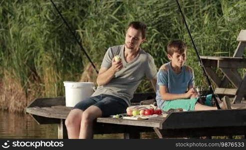 Positive stylish dad and teenage boy having a picnic on a wooden pontoon in summer sunlight while fishing together by the lake. Joyful father and son enjoying a meal and communicating with each other. Slow motion.