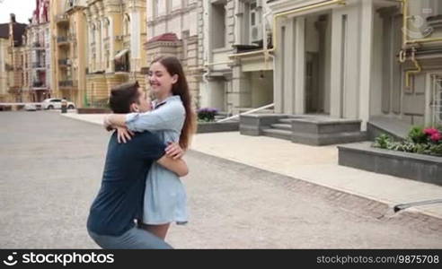 Positive young couple in love spinning around during a romantic date in the city street. Happy young woman embracing boyfriend's neck delicately while man holding her waist tightly in hands and whirling beloved girl. Slow motion. Stabilized shot.