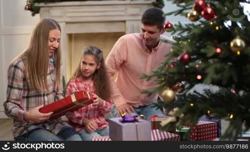 Positive young family of three exchanging gifts near Christmas tree while spending winter holidays at home. Joyful parents and excited adorable daughter sitting near Christmas tree and exchanging different presents in festive decorated room. Dolly shot.