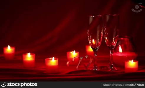 Pouring champagne in glasses at Valentine's Day celebration, still life with candles, roses, and hearts on red silk