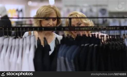 Pretty senior female friends doing shopping in apparel boutique store. Beautiful women buying clothes during shopping in fashion garments shop. Shopaholic females selecting clothing behind the rack with different apparels hanging on hangers.