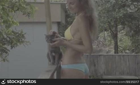 Pretty young woman wearing bikini standing and playing with cat on the terrace in slow motion
