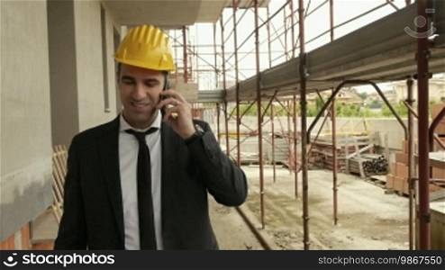 Profession, business, and telecommunications. Architect talking on a cell phone and walking on a construction site under building scaffolding. Steadicam shot.