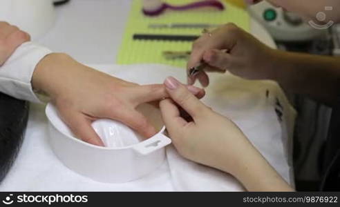 Professional manicurist doing cuticle cleaning on female fingers in beauty salon. Close up nail beautician's hands removing cuticles with professional nail tool. Manicurist doing cuticle cleaning on female finger with metal clippers.