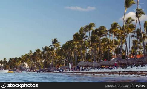 PUNTA CANA, DOMINICAN REPUBLIC - NOVEMBER 14, 2014: People walking on the beach bordered with high palms, touristic boats sailing to the shore