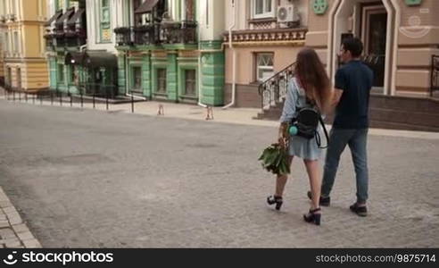 Rear view of loving couple stepping down cobblestone sidewalk during romantic date. Attractive woman carrying bunch of flowers holding hand of boyfriend walking down the street while enjoying time together outdoors. Slow motion. Steadicam stabilized shot.