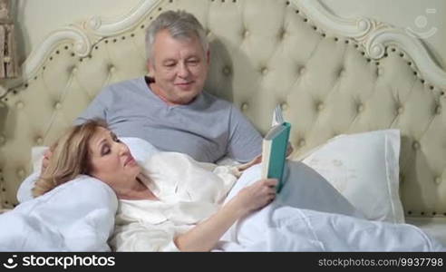 Relaxed elderly woman lying down on her back with her head on husband's lap and reading a book in bed while retired man caressing her hair. Mature couple enjoying time together in bedroom and lounging.