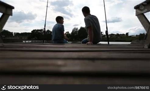 Relaxed father and teenage son bonding and communicating with each other while sitting on a wooden pier and fishing on a calm pond. Back view. Low angle view. Positive dad and his boy angling together on the lake over a cloudy sky background.