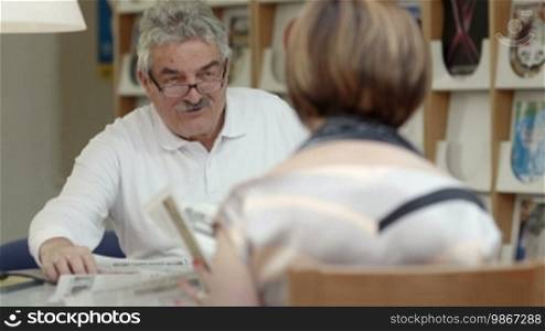 Retired senior people, old man reading paper with wife in library. Dolly shot