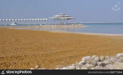 Scene of the resort on Dead Sea. Beach and water view with few people relaxing under the shed in distance