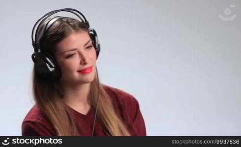 Seductive young brunette woman wearing red lipstick and listening to music in headphones. Sexy pretty girl with playful expression, twisting long hair around her finger and looking at the camera teasingly while enjoying favorite track in earphones.