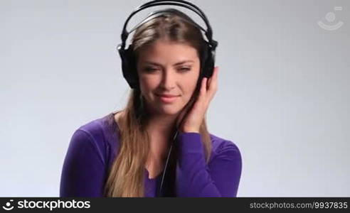 Seductive young brunette woman with long hair listening track on mp3 player on white. Happy smiling girl in earphones enjoying music, touching one earcup, hearing the song fading, slowly taking off headphones and hanging them around her neck.