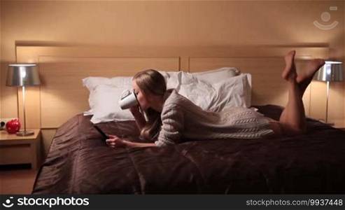 Seductive young woman wearing cardigan and tan stockings lying on the bed in luxury style bedroom using digital tablet while drinking hot fresh cup of coffee. Charming sexy blond female relaxing at home after work.