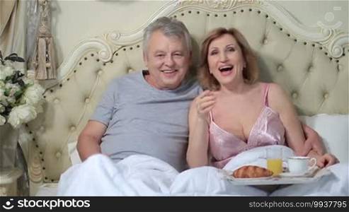 Senior couple lying in bed in the morning and watching television at home. Smiling married aged couple enjoying comedy show on TV while lounging in the bed.