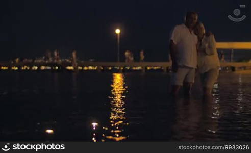 Senior couple standing barefoot in sea near the pier at night and looking at candles floating on dark rippling water with reflection of golden lantern light