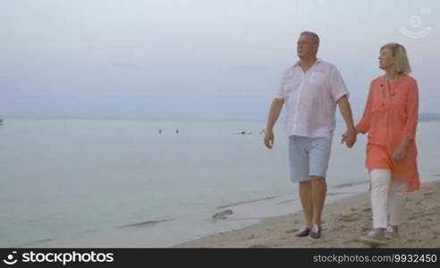 Senior family couple having a quiet outdoor walk in the evening. They are walking hand in hand along the calm sea and talking