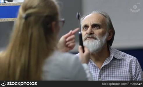 Senior male patient with beard checking nervous system by professional neurologist in medical office. Elderly man following test hammer during medical exam in hospital. Female neurologist testing reflexes of patient's eyes using test hammer.