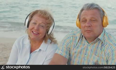 Senior man and woman listening to music in wireless headphones by the sea. They enjoying songs as dancing with favorite rhythms. Relaxing outdoor