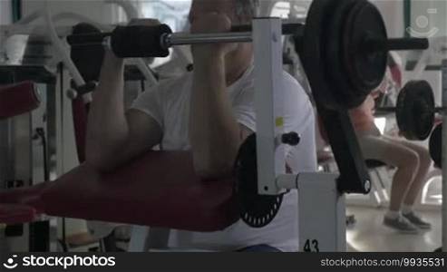 Senior man exercising on bicep curl machine, woman training on chest press machine in background
