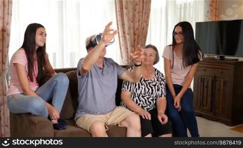 Senior man in virtual reality headset or 3D glasses having fun with his wife and granddaughters. Senior man wearing virtual reality glasses at home. People having fun with new technology concept. Slow motion