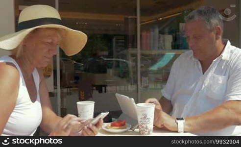 Senior people in an outdoor cafe. Everyone is captured with electronics. Man using a pad and smartwatch, woman typing on a smartphone
