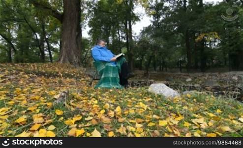Senior woman enjoying her retirement in a chair under an autumn tree, reading a book. Dolly shot, wide angle, side view.