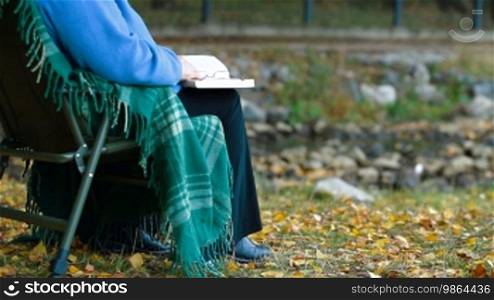 Senior woman enjoying retirement with a book at autumn park by the river. Unrecognizable person, side view.