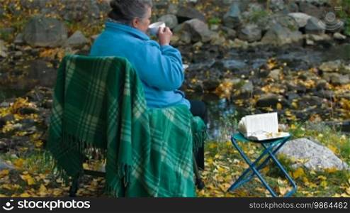 Senior woman holding white coffee cup while sitting at autumn park