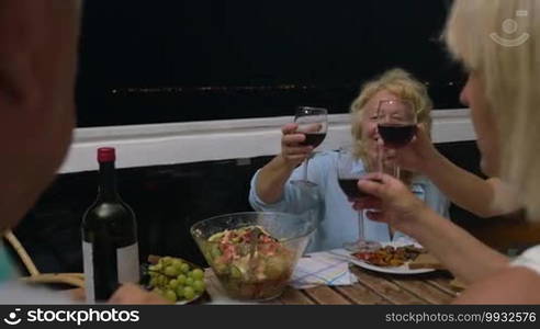 Senior woman proposing a toast during late dinner in outdoor balcony. Family members clanging glasses and having wine. City lights can be seen in distance