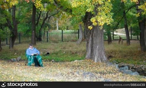 Senior woman sitting outdoors in chair under autumn tree, reading book. Front View.