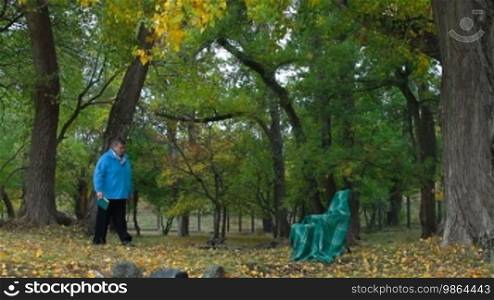 Senior woman sitting outdoors in chair under autumn tree, reading book. Side View.