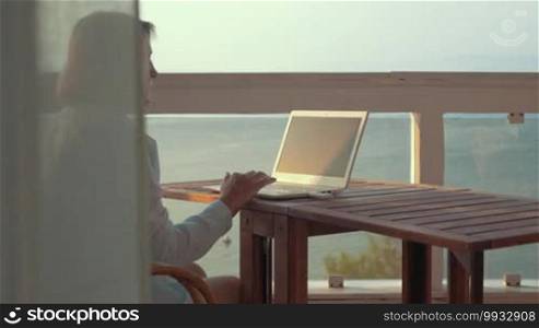 Senior woman using a laptop sitting at the table on an outdoor balcony with a view of the sea. She is surfing online using a mobile internet device connected through USB.