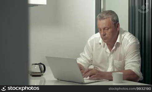 Serious adult man working on laptop while drinking coffee in kitchen. Man wearing glasses.