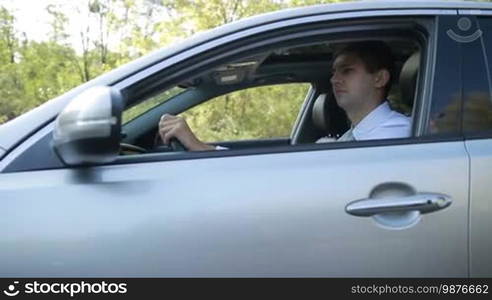 Serious young businessman driving car on rural road in summer during business trip. Confident man enjoying travel on countryside in modern vehicle. Side view. Slow motion. Steadicam stabilized shot.