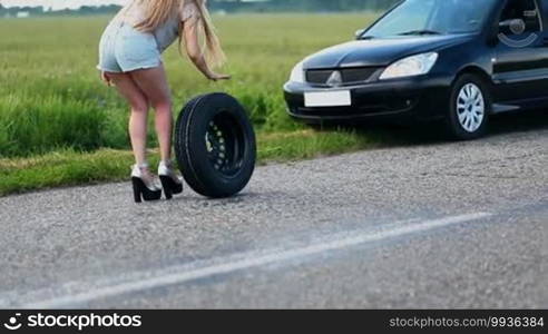 Sexually dressed woman is rolling spare wheel on rural road to change flat tire