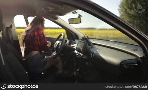 Sexy female driver in shorts and plaid shirt getting into parked car, fastening seatbelt and going on summer road trip vacation with awe landscape on background. Seductive woman traveling by car on rural road on sunny day. Slow motion. Stabilized shot.