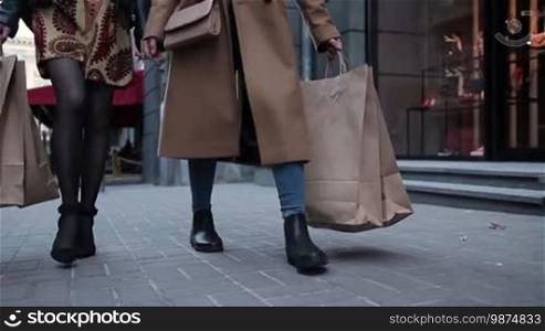 Shopping day. Close-up of young women carrying shopping bags while walking along the shopping street. Slow motion. Midsection of girls holding shopping bags stepping on cobblestone sidewalk near city stores.