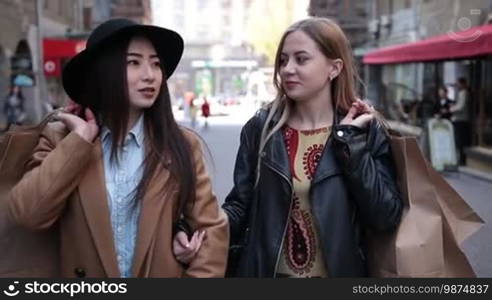 Shopping girls. Two multiracial young girlfriends with shopping bags over shoulders having fun while buying clothes in shopping mall. Slow motion. Beautiful elegant Asian woman and Caucasian female friends shopping together.