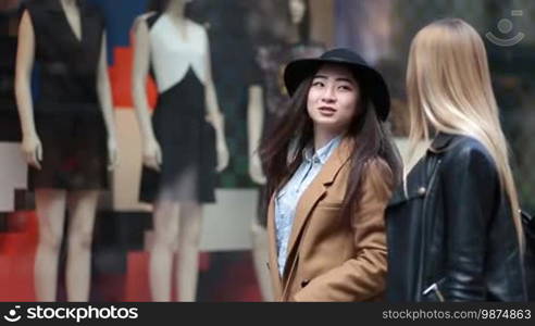 Shopping young women looking at clothing store window outside shop. Multiethnic girlfriends standing in front of expensive boutique store display window and shocked by price tags. Blurred boutique display window with mannequins on background.