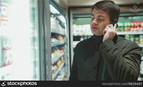 Shot of a man in a food shop. He's talking on the phone and choosing a beverage in the refrigerator.