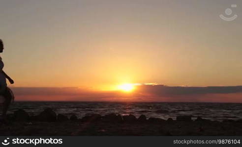 Side view of affectionate couple in love approaching each other and standing face to face holding hands on the beach in rays of orange setting sun. Romantic couple expressing feelings, love and tenderness at seaside against setting sun background.