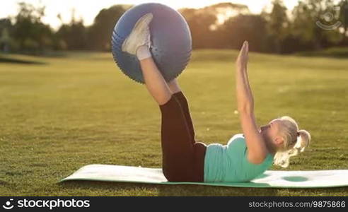 Side view of an attractive sporty senior woman in sports clothes training with a blue fitball while lying on her back on an exercise mat in the park at sunset. A charming adult fit blonde female doing abdominal crunches with a fitball in the fresh air.